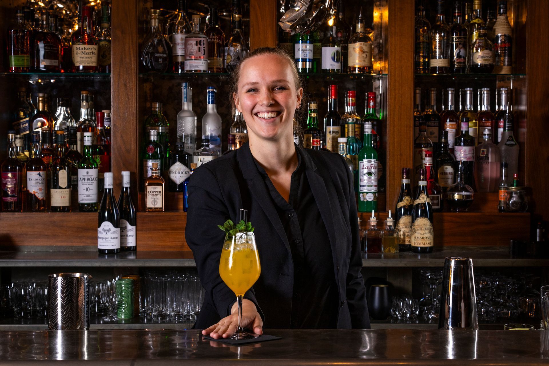 Bartender smiles, presenting a bright orange cocktail at a bar with many liquor bottles.