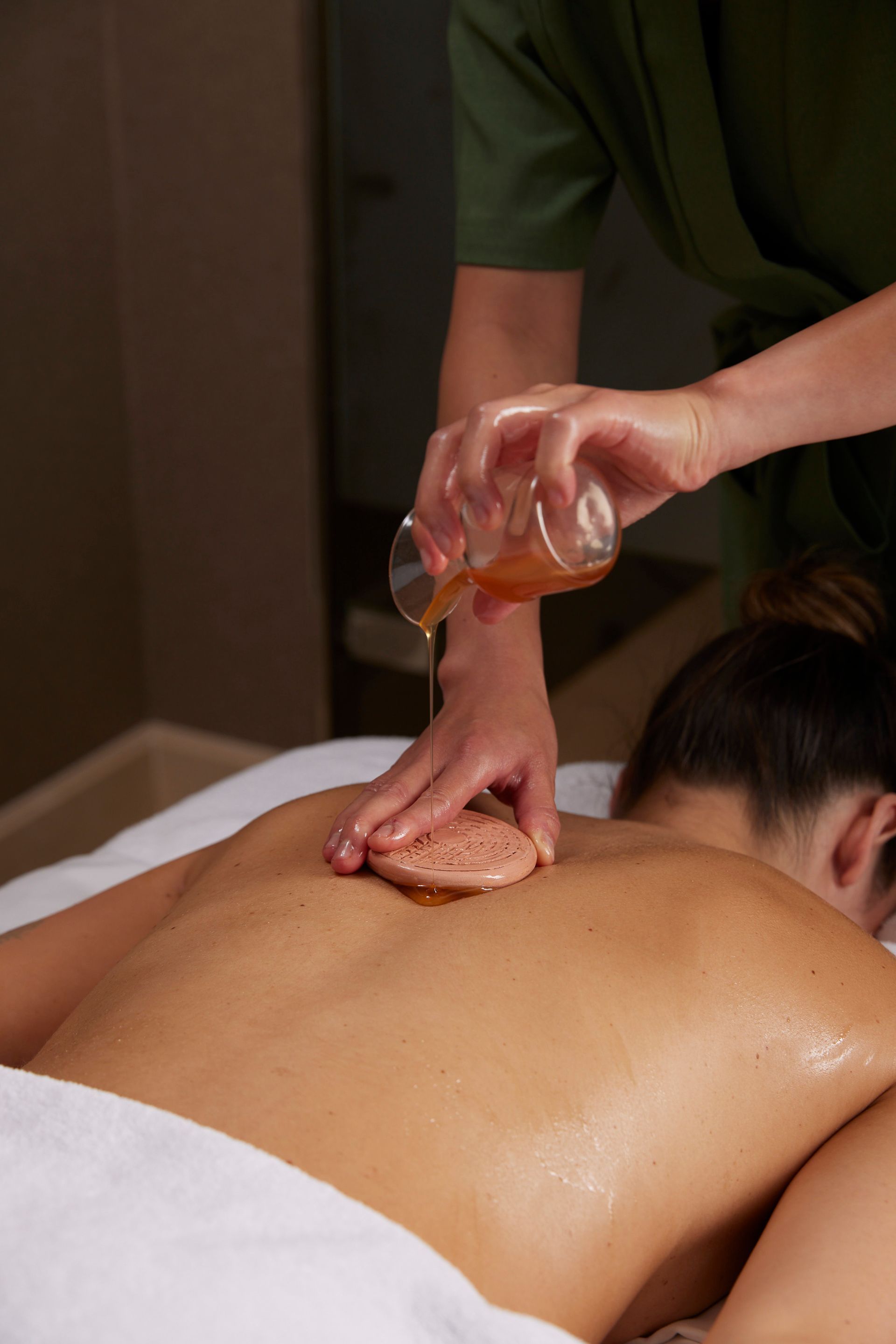 Person receiving back massage, oil being poured on sponge, spa setting.