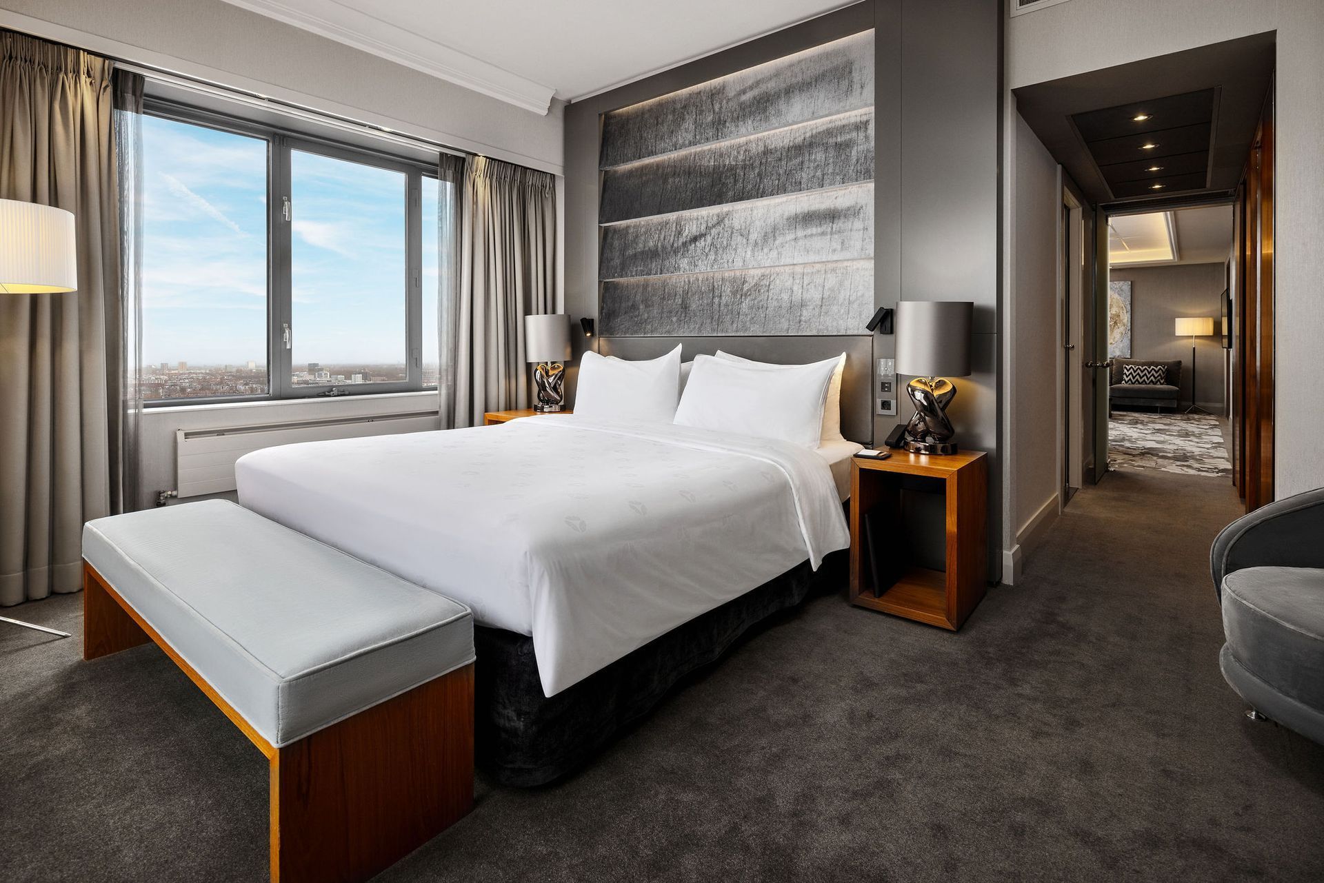 Hotel bedroom with a king bed, bench, window, and hallway entrance. Gray walls and carpet.