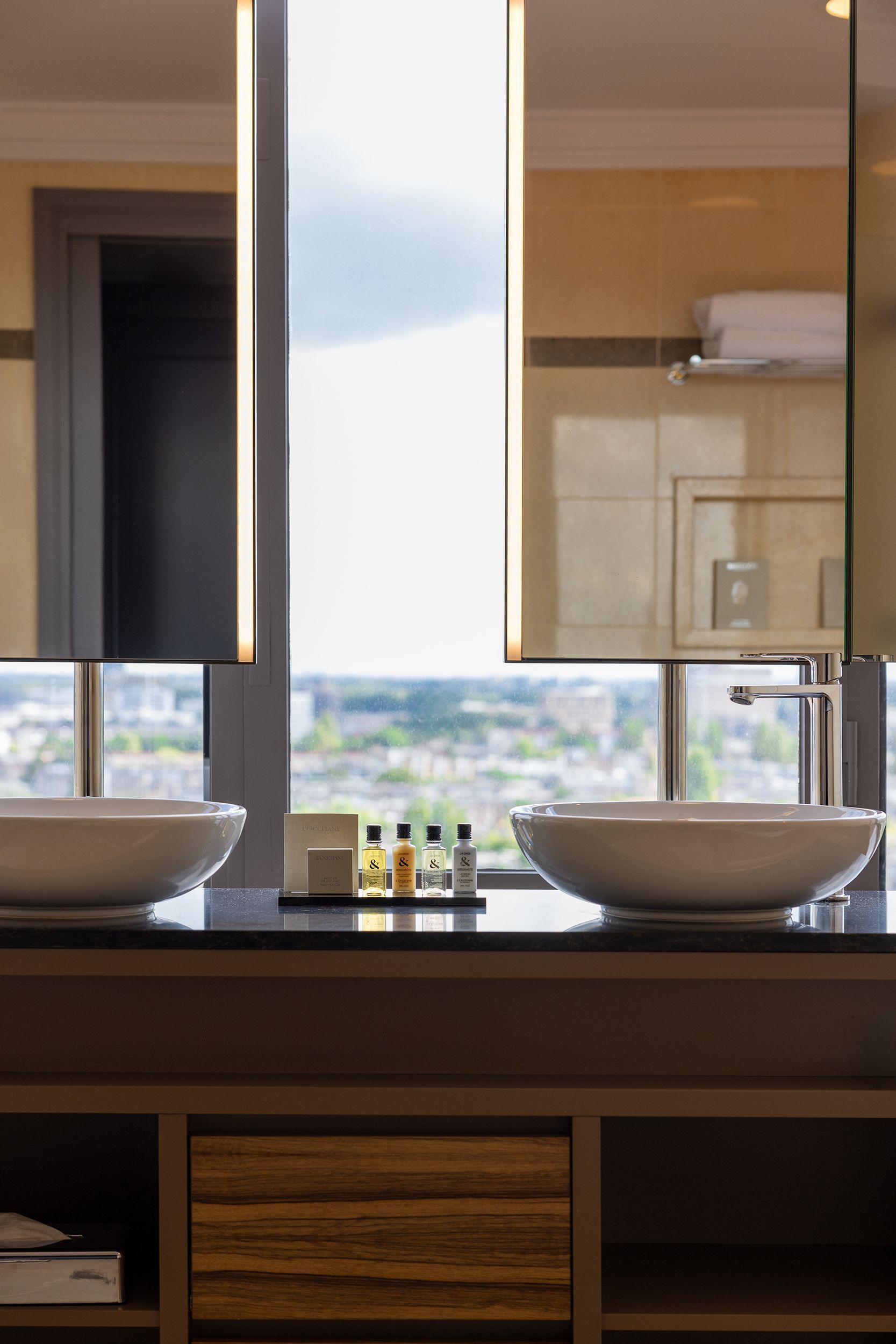 Bathroom with two sinks, mirrors, toiletries, and a city view.
