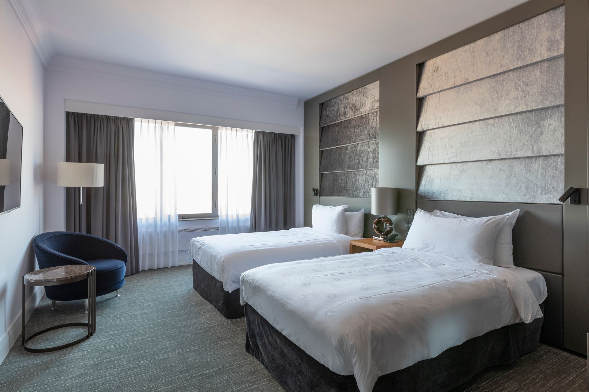 Hotel room with two twin beds, small table, chair, window with drapes, and a lampshade. Gray and white color scheme.