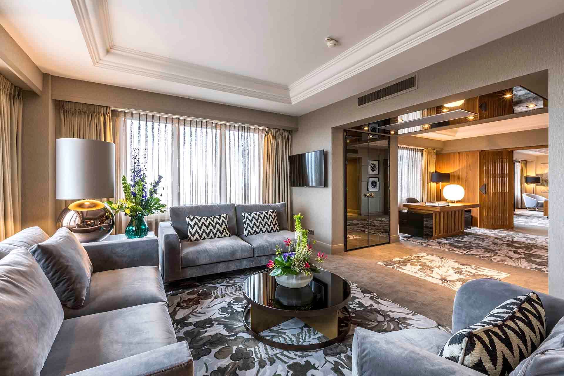 Luxurious hotel suite with gray couches, floral rug, and mirrored wall.