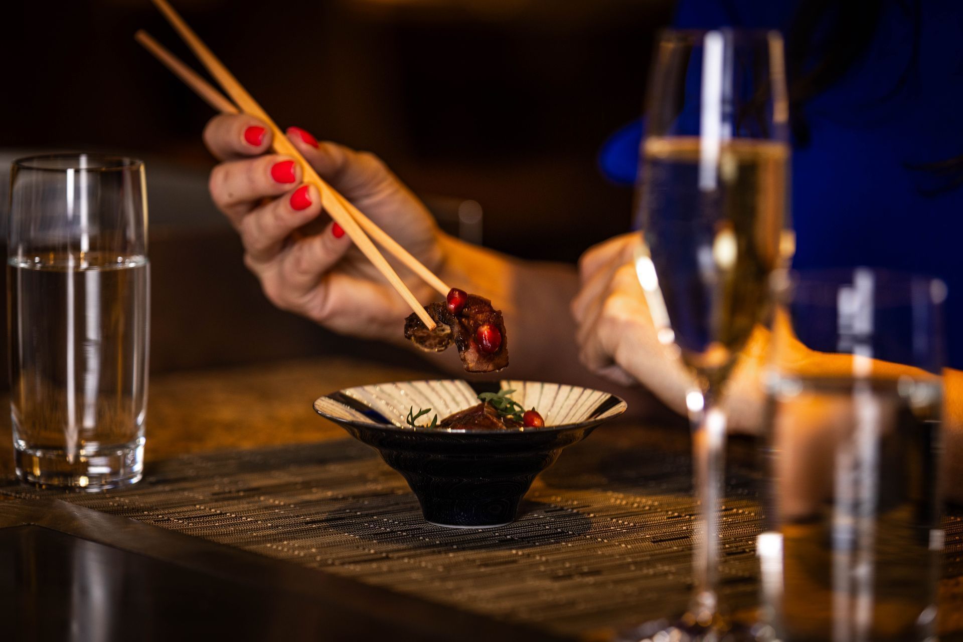 Person uses chopsticks to eat food from a small bowl. Champagne and water glasses are nearby.