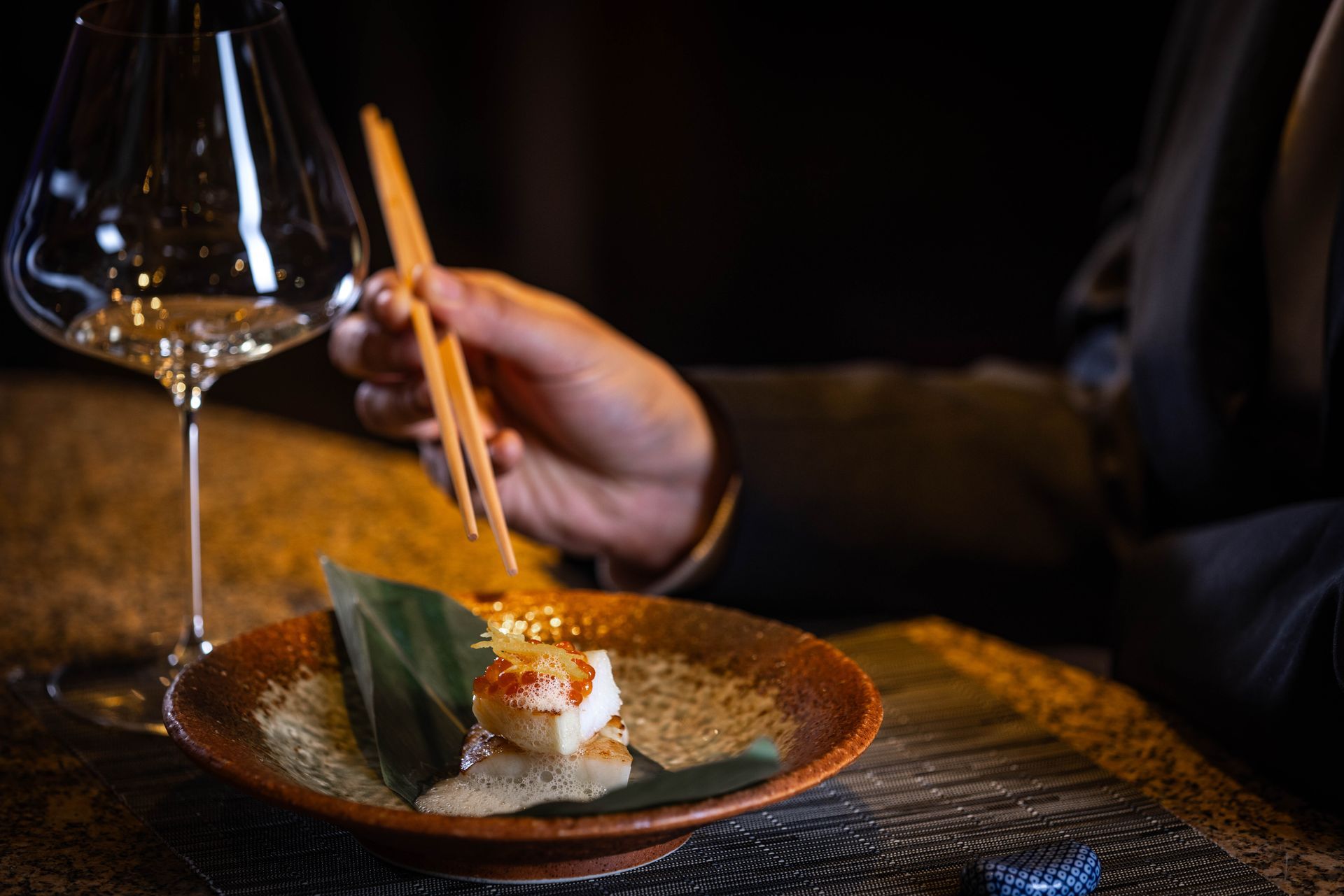 Person using chopsticks to eat seafood from a small, decorative bowl next to a wine glass.