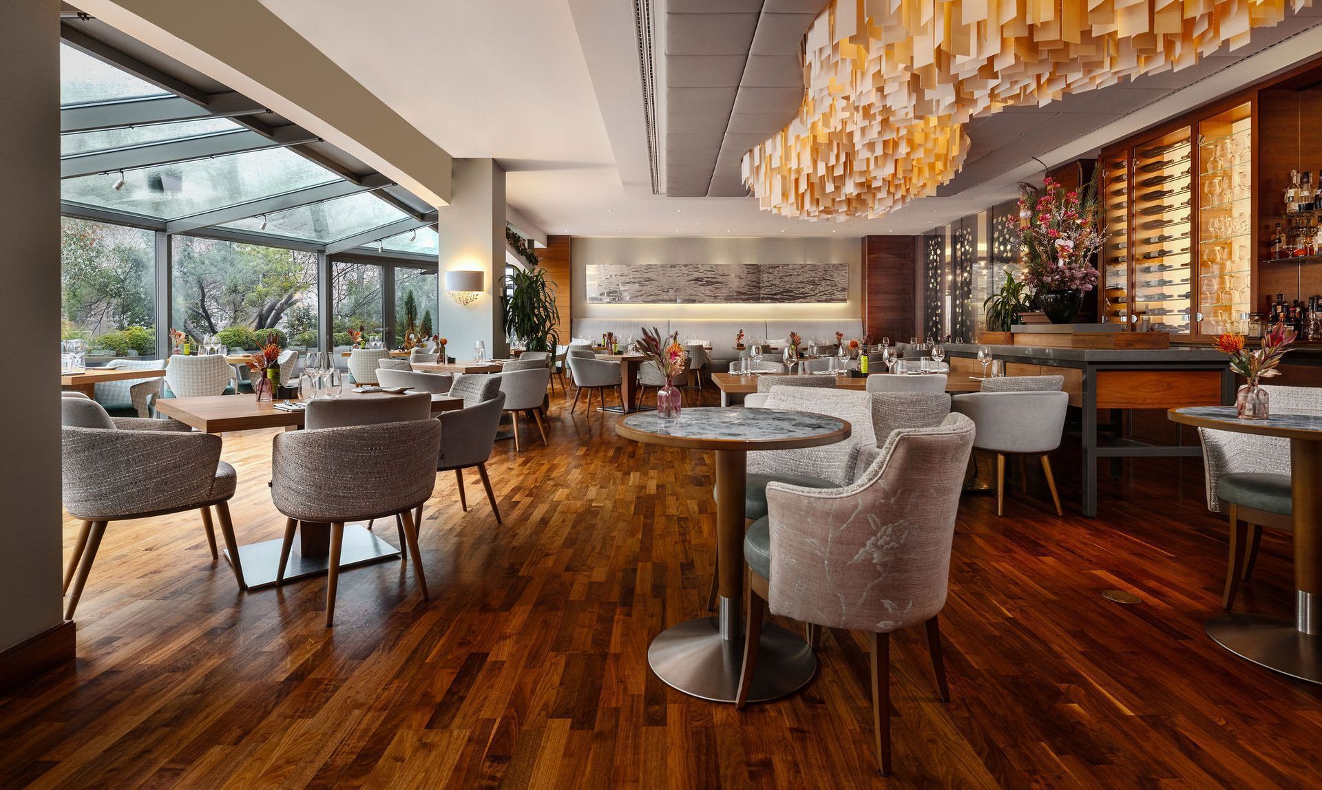 Restaurant interior with tables and chairs on a wooden floor, a glass ceiling, and decorative chandelier.