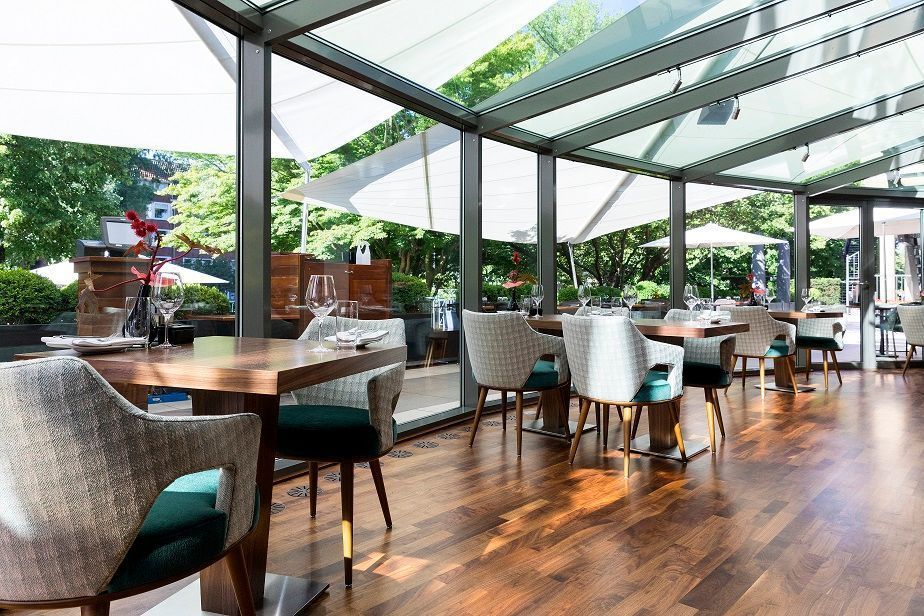 Restaurant interior with wooden floor, tables, chairs, and large windows overlooking trees.