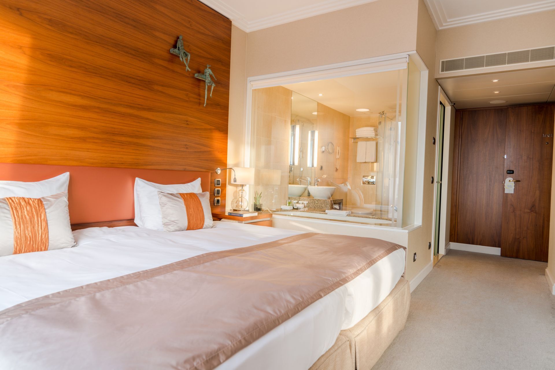 Hotel bedroom with king-size bed, ensuite bathroom visible through a glass wall, and wood-paneled feature wall.