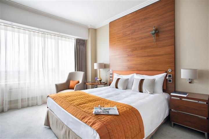 Hotel room with a bed, chair, and large wooden headboard. Orange accents contrast the neutral tones.