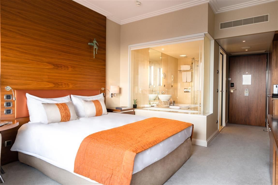 Hotel room with king bed, open bathroom, wood paneling, orange accents.