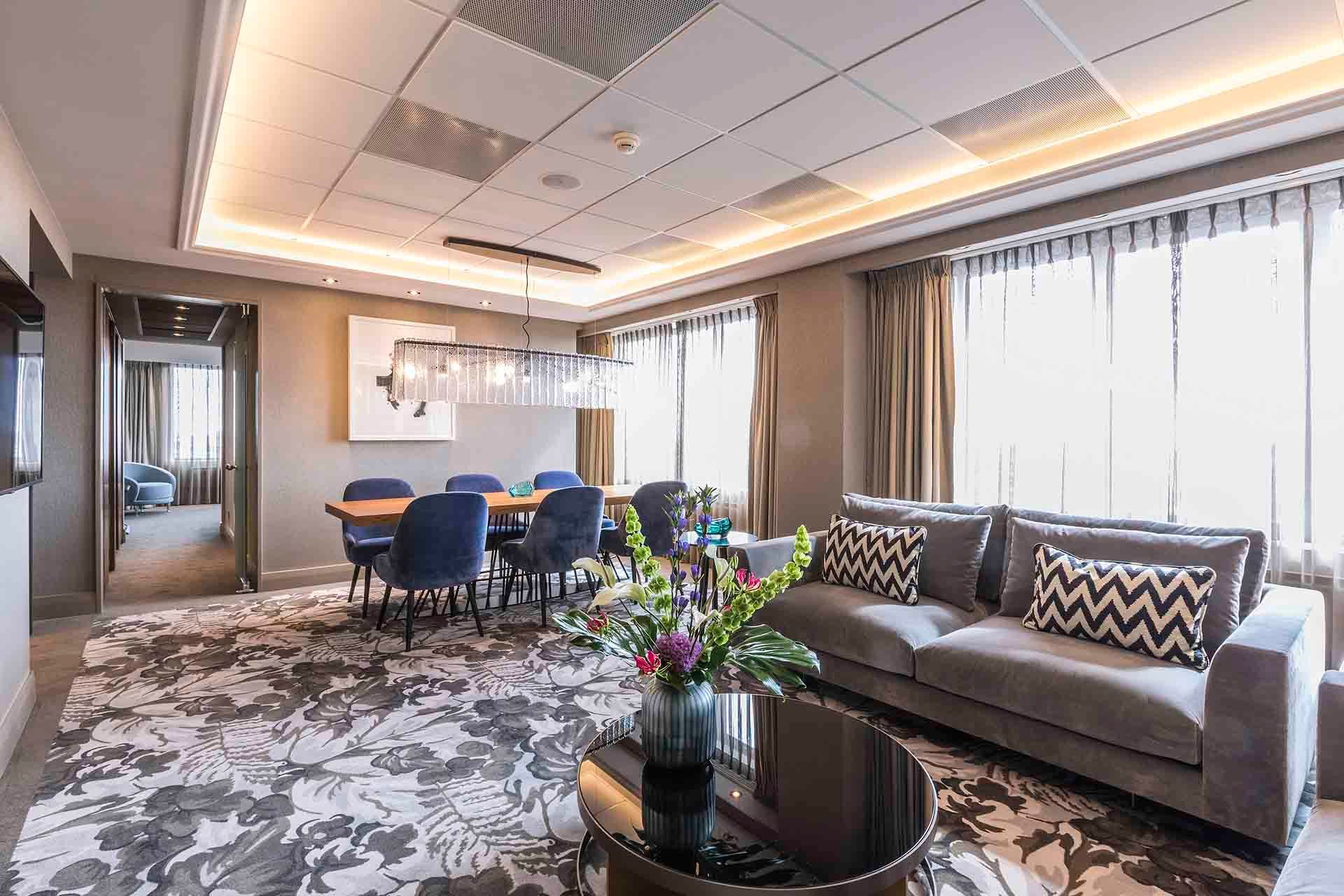 A luxurious hotel suite with a dining area, living room with gray sofa, and a floral rug.