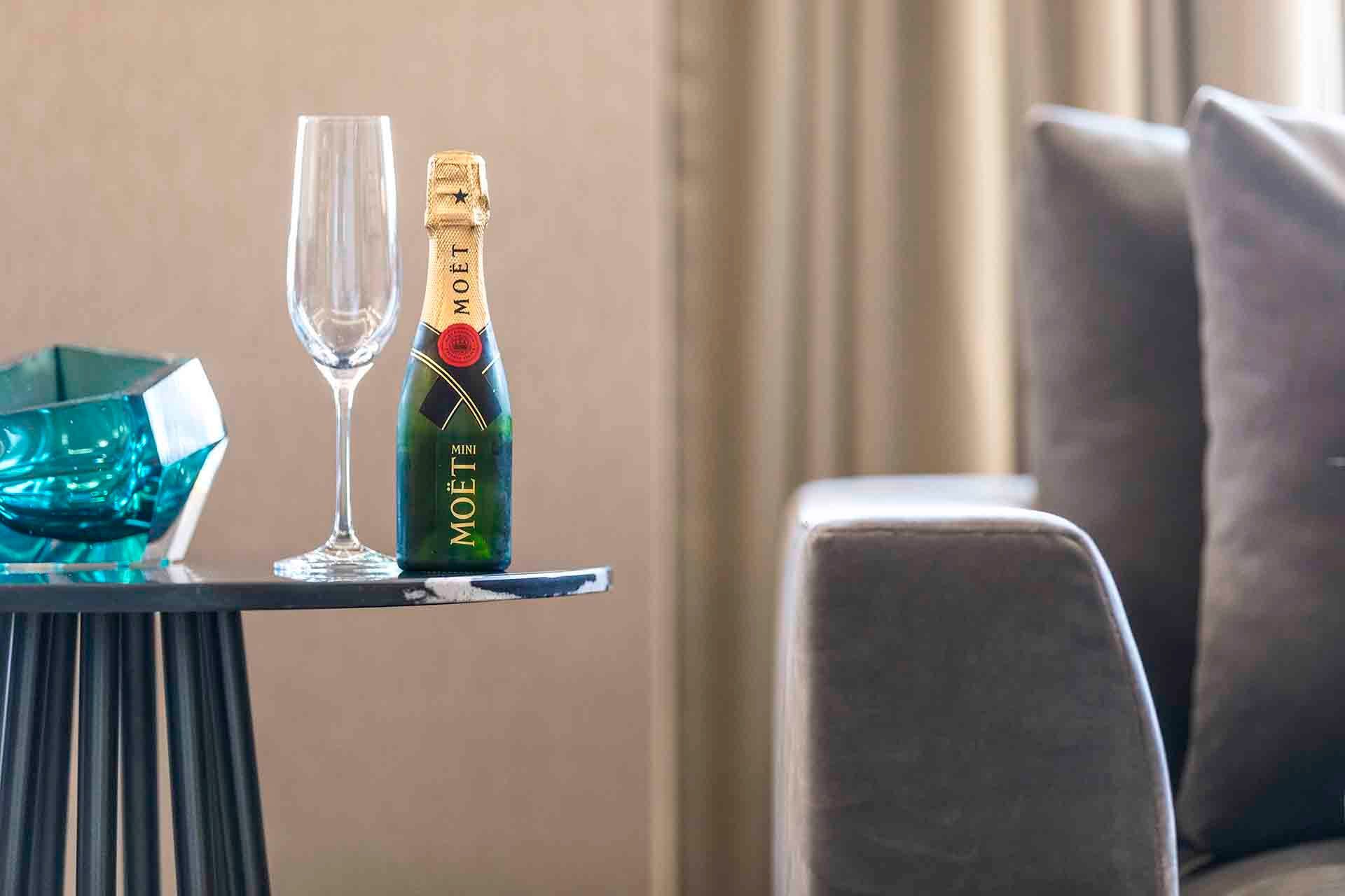 A bottle of Moët champagne, a glass, and a blue bowl sit on a black side table next to a sofa.