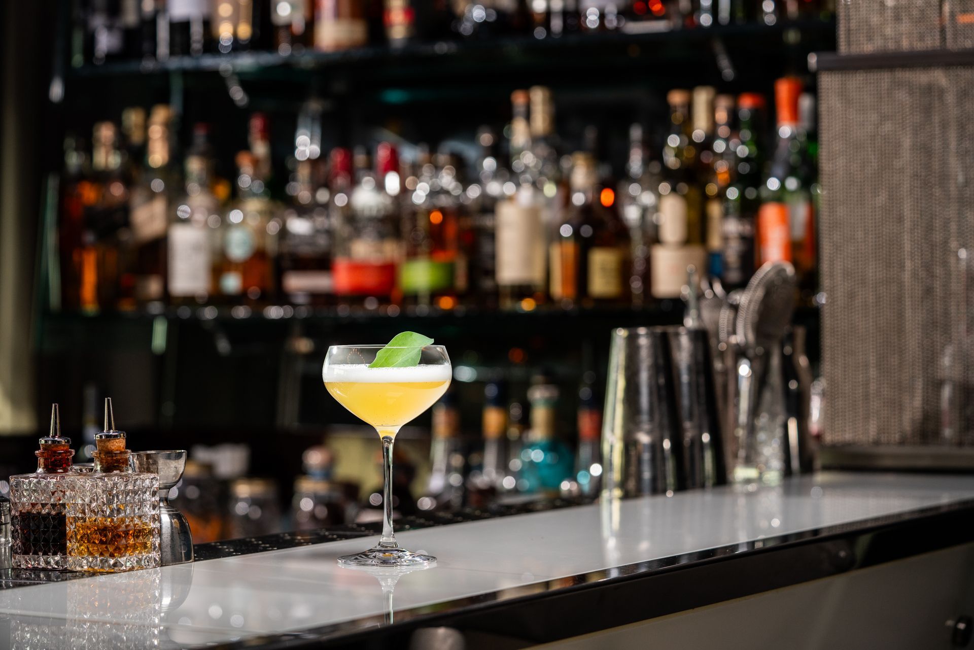Cocktail on bar, basil garnish. Bottles on shelves in the background.