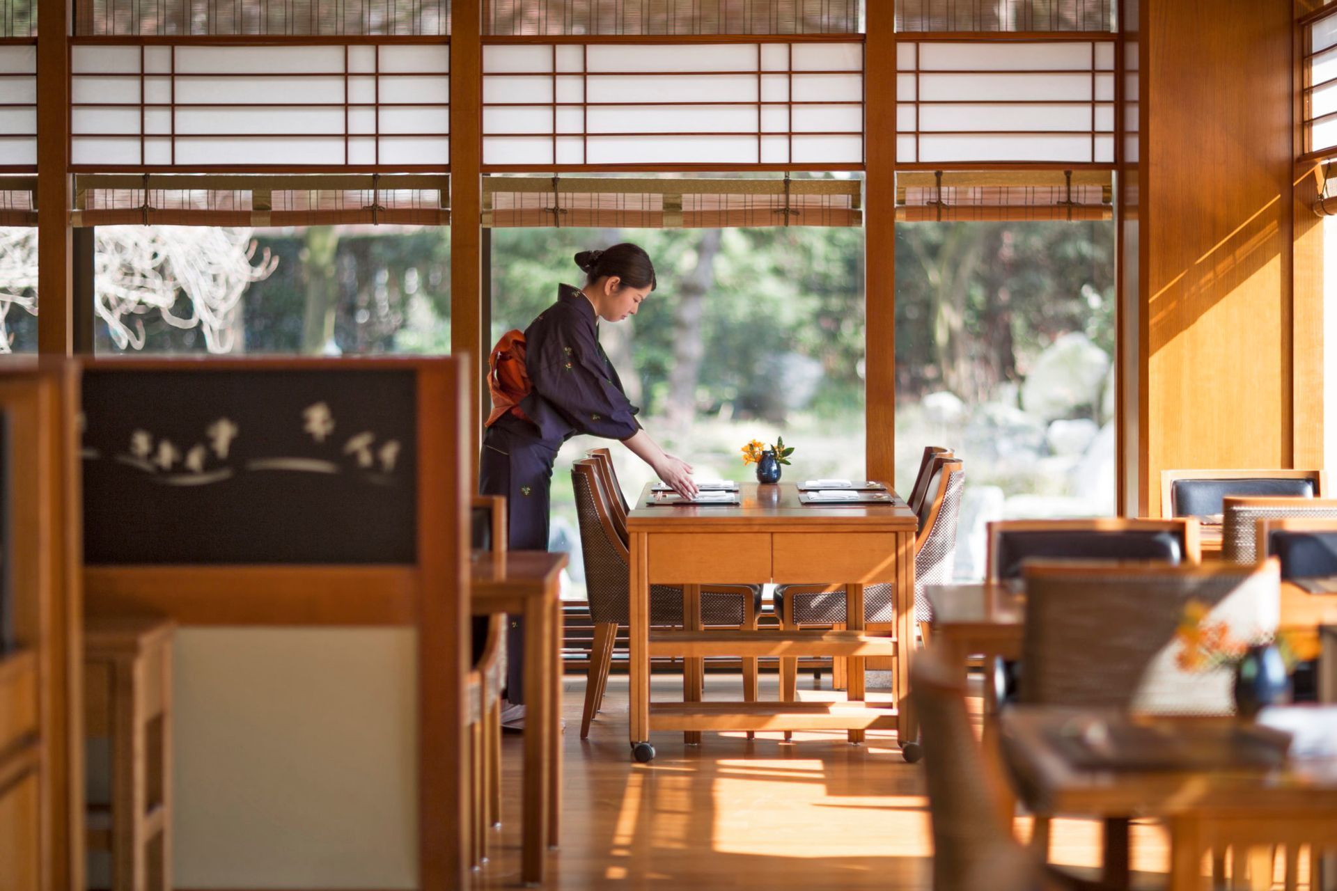 Woman setting a table in a Japanese-style restaurant with wooden decor and large windows.