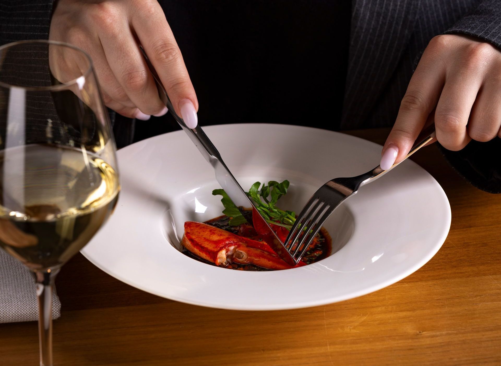 Person using knife and fork to eat a plate of food, with a glass of wine on a wooden table.