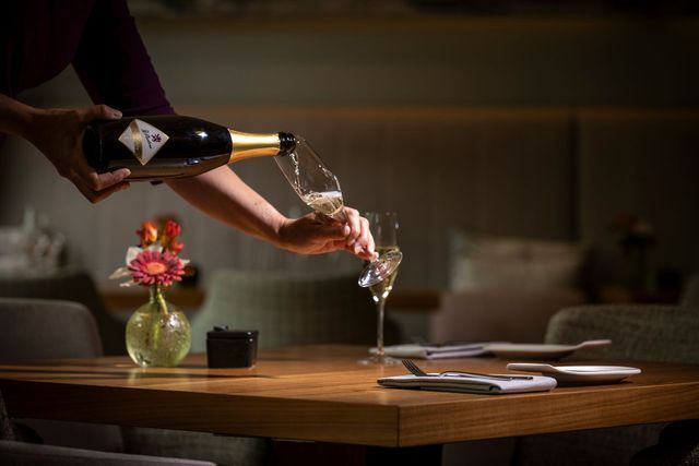 Person pouring champagne into two glasses at a table, floral centerpiece, plates, and a dark, intimate setting.