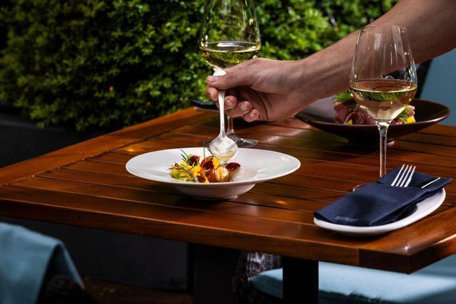 Person placing food on a plate at a table set for dining, with wine glasses and napkin.