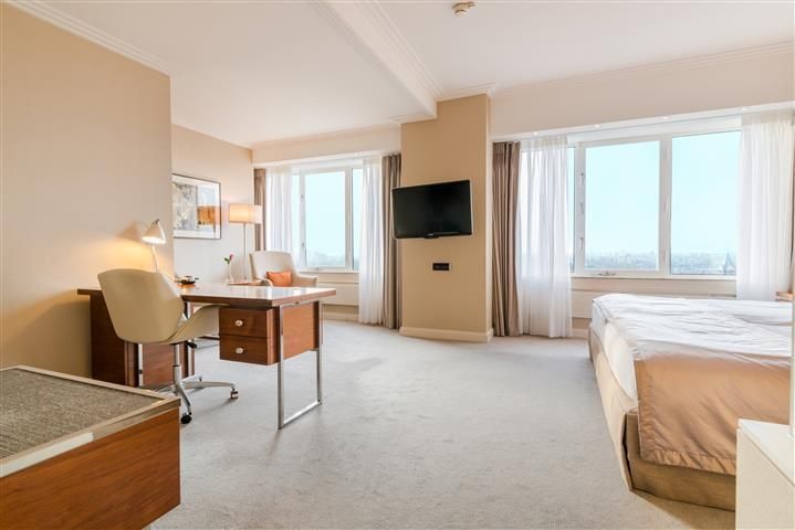 Hotel room with a desk, bed, TV, and large windows with cityscape views. Beige walls, carpet.