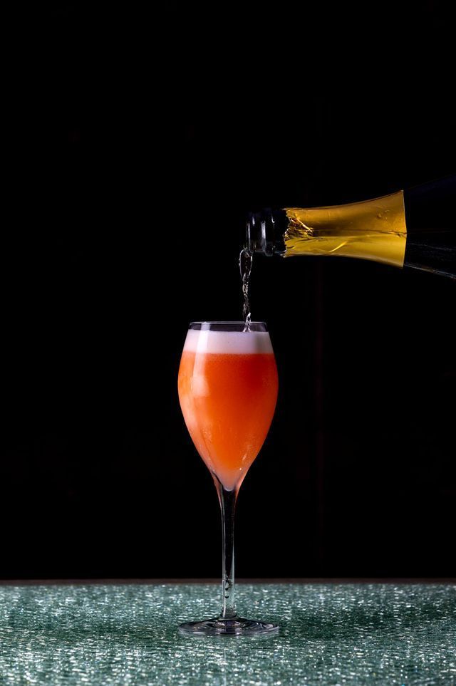 Champagne being poured into a tall glass with orange liquid, on a sparkly surface, against a black background.