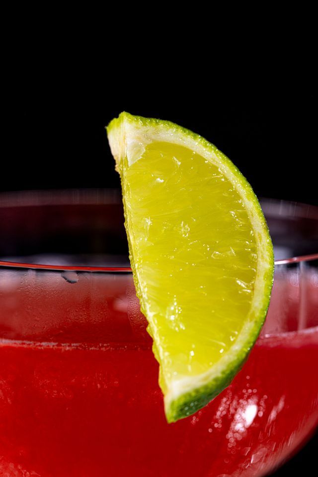 Close-up of a red cocktail in a glass, garnished with a lime wedge against a black background.