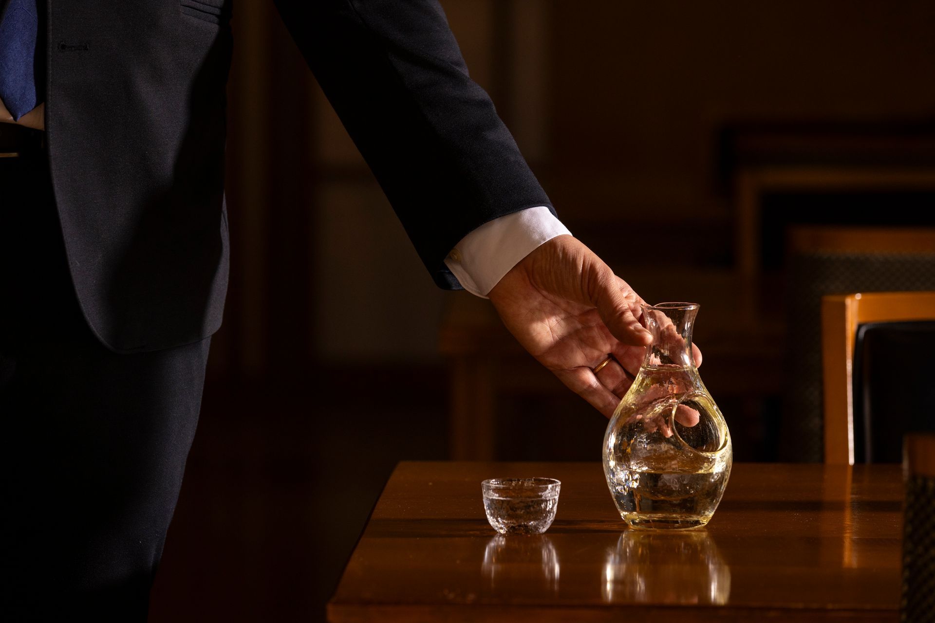A person in a suit pouring liquid from a glass decanter into a small cup on a table.