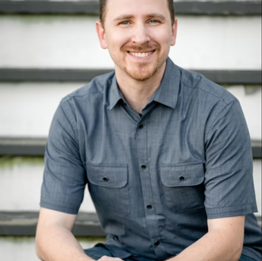 Man with a beard in a blue button-down shirt smiles, sitting on outdoor stairs.