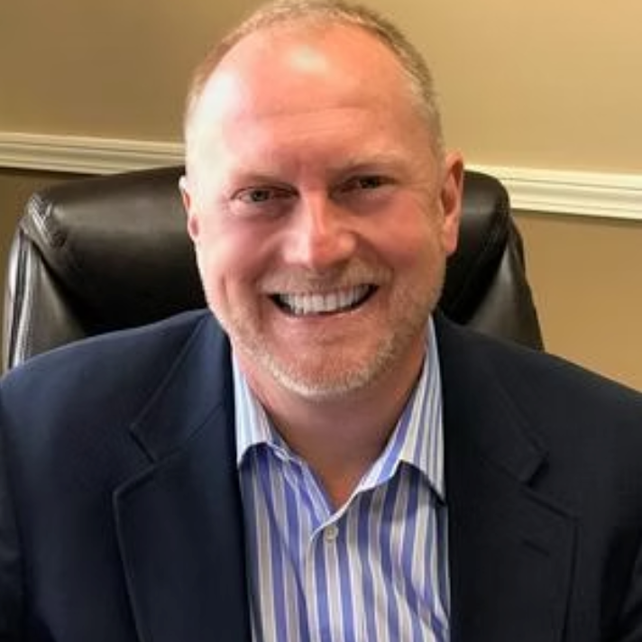 Man with short hair, smiling, in a blue blazer and striped shirt. Sitting at a desk.