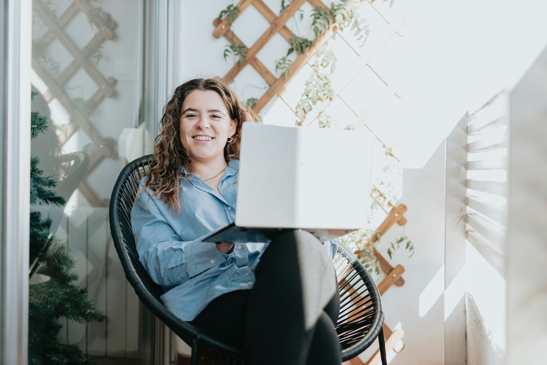 Woman in a chair on a balcony smiles while using a laptop.
