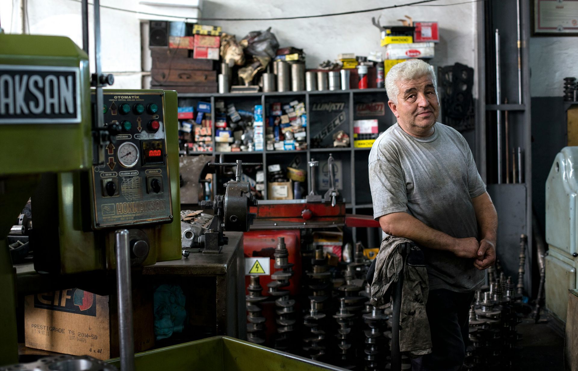 Man in a garage with machinery, smiling, leaning. Shelves and tools in the background.