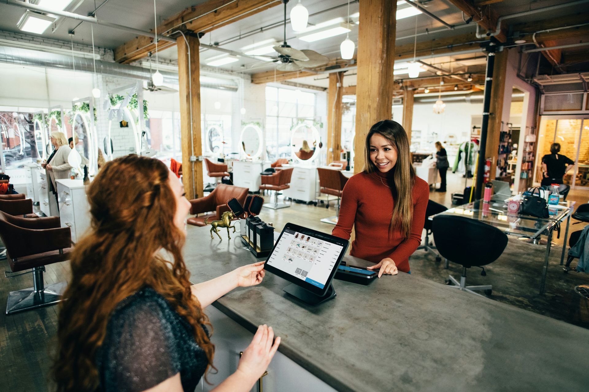 A woman at a salon counter paying with a tablet, with a smiling stylist.