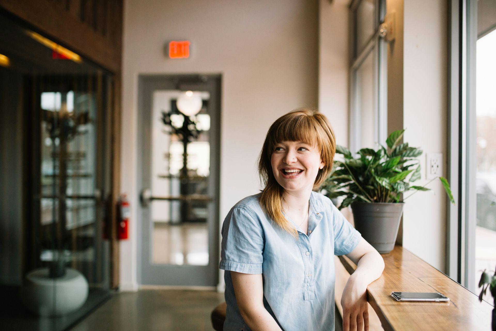 Woman with red hair smiles by a window, resting arm on a wooden ledge, with potted plant beside her.