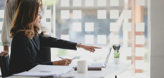 Two businesswomen collaborate at a desk, one pointing at a laptop screen.