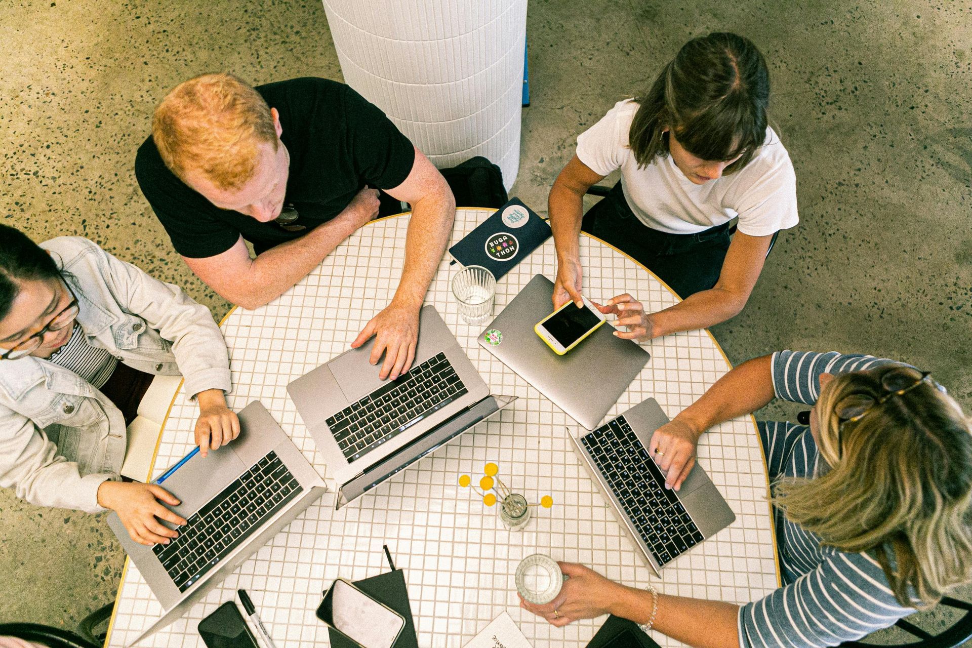 Four people at a round table working on laptops and phones, possibly in a cafe.
