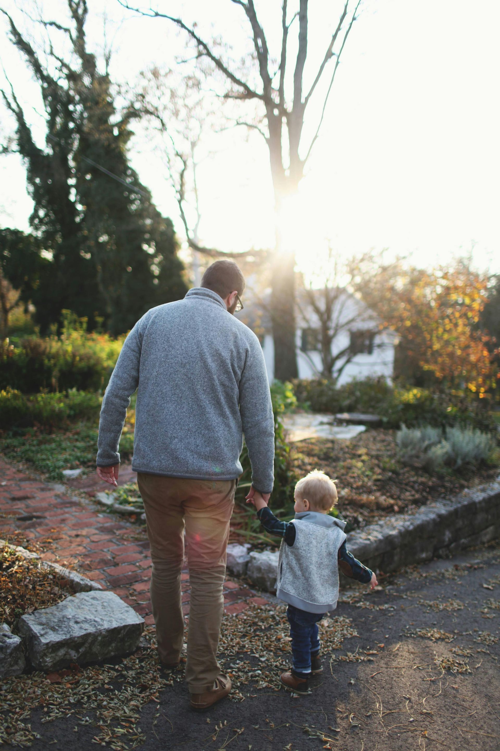 Father and toddler walk hand-in-hand on a path, sun shining, autumnal scene.