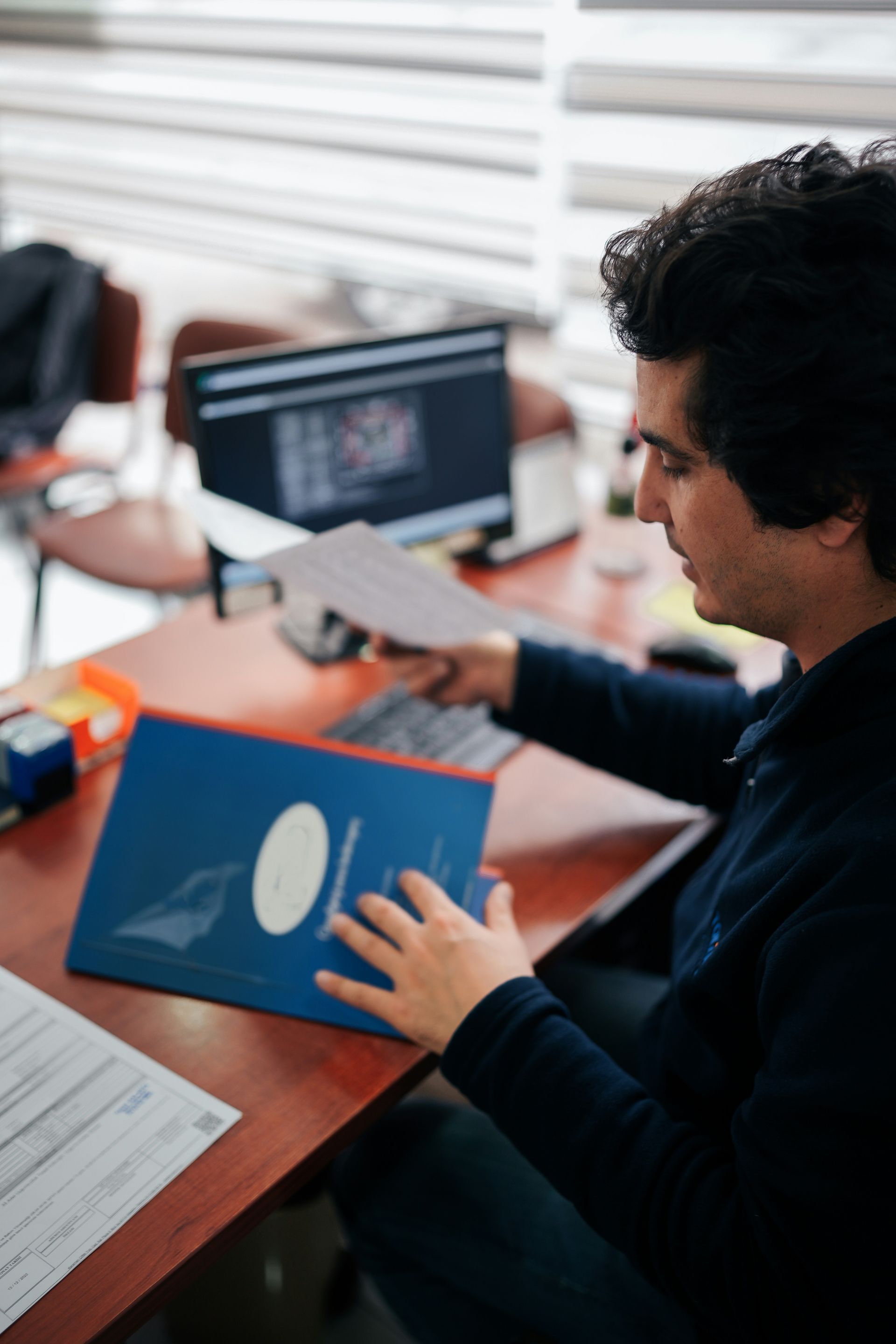 Man at desk holding papers, looking at a blue binder. Computer in background.