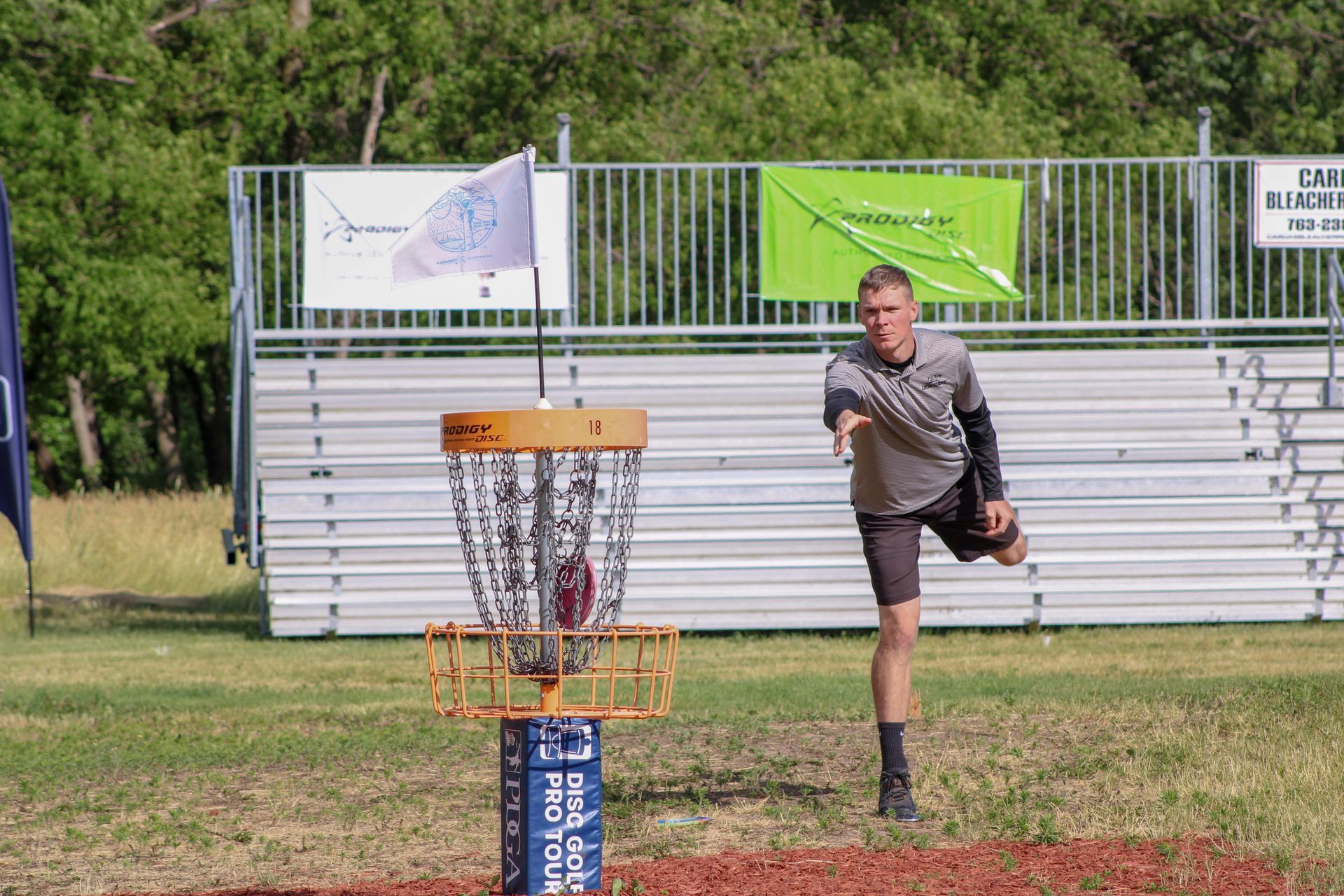 Man throwing a disc at a basket on a disc golf course; stands near an empty grandstand.