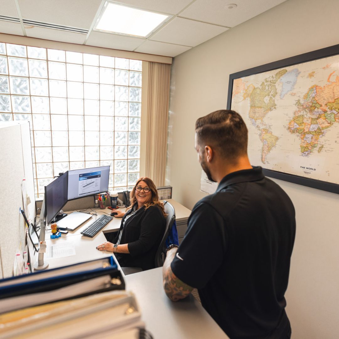 A man is standing next to a woman sitting at a desk in an office.