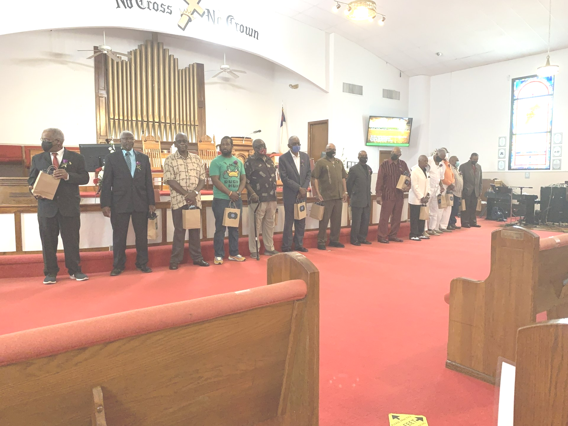 People standing in a church, facing forward. Several men in a line. A red carpet and wooden pews.