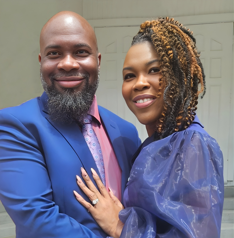 A smiling couple poses for a portrait, with the person on the right wearing a blue top and resting a hand on the other.