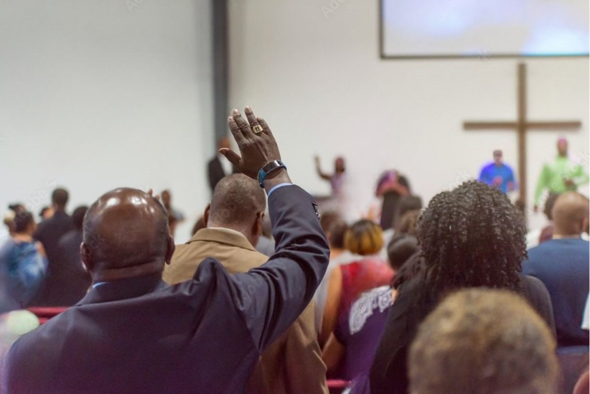 People raising hands in a church, focused on a man in a blue suit. Cross and screen in the background.