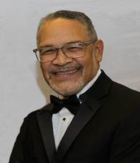 Man in a tuxedo with glasses and bow tie, smiling.