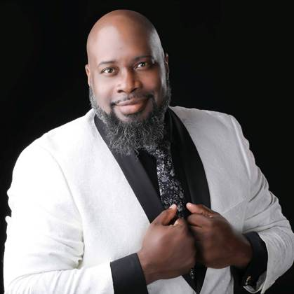 Clergyman holding an open book, smiling, wearing a black robe with crosses, against a sky-blue background.