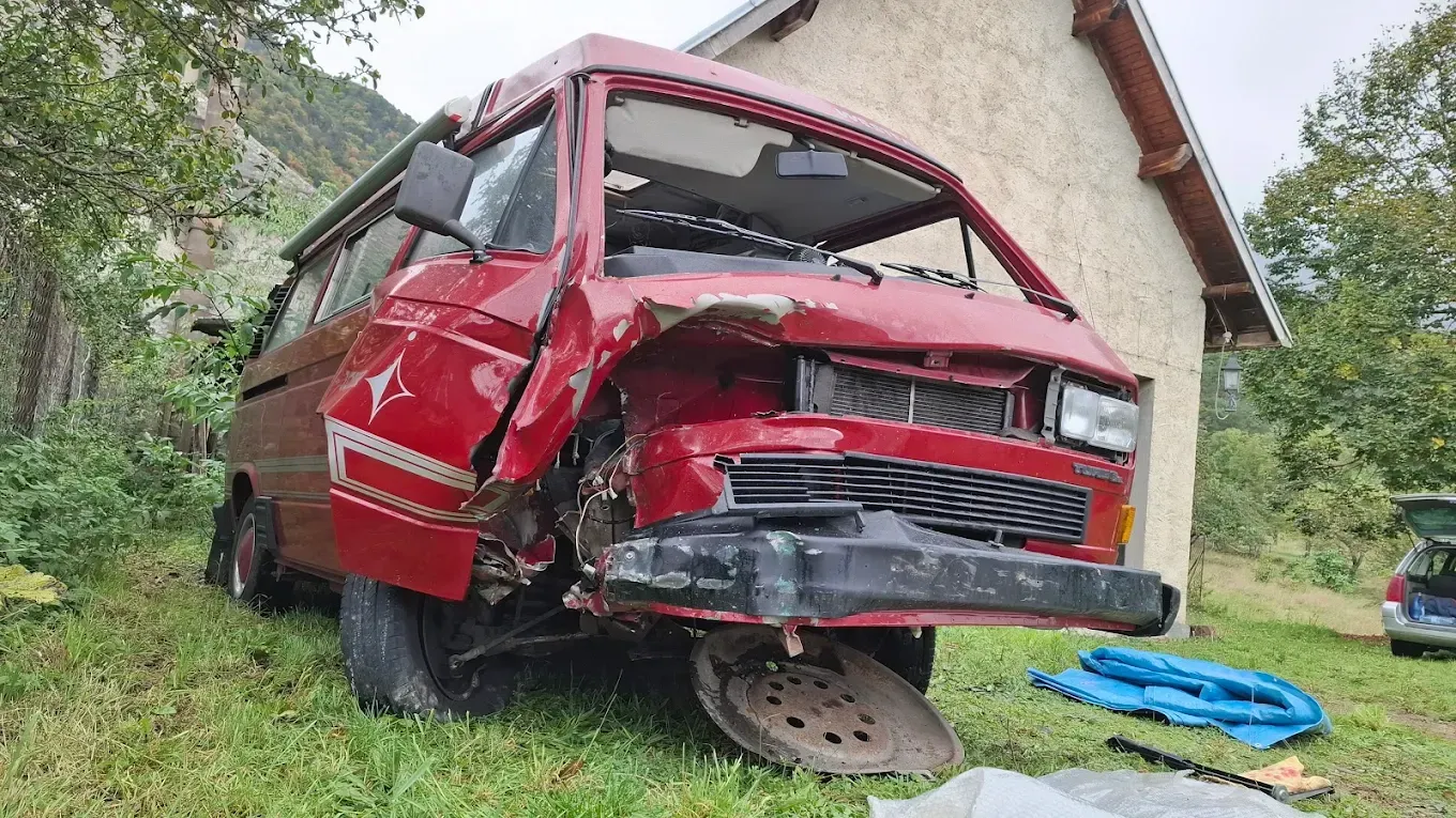 Une camionnette rouge fortement endommagée gît sur l'herbe, à côté d'un bâtiment.