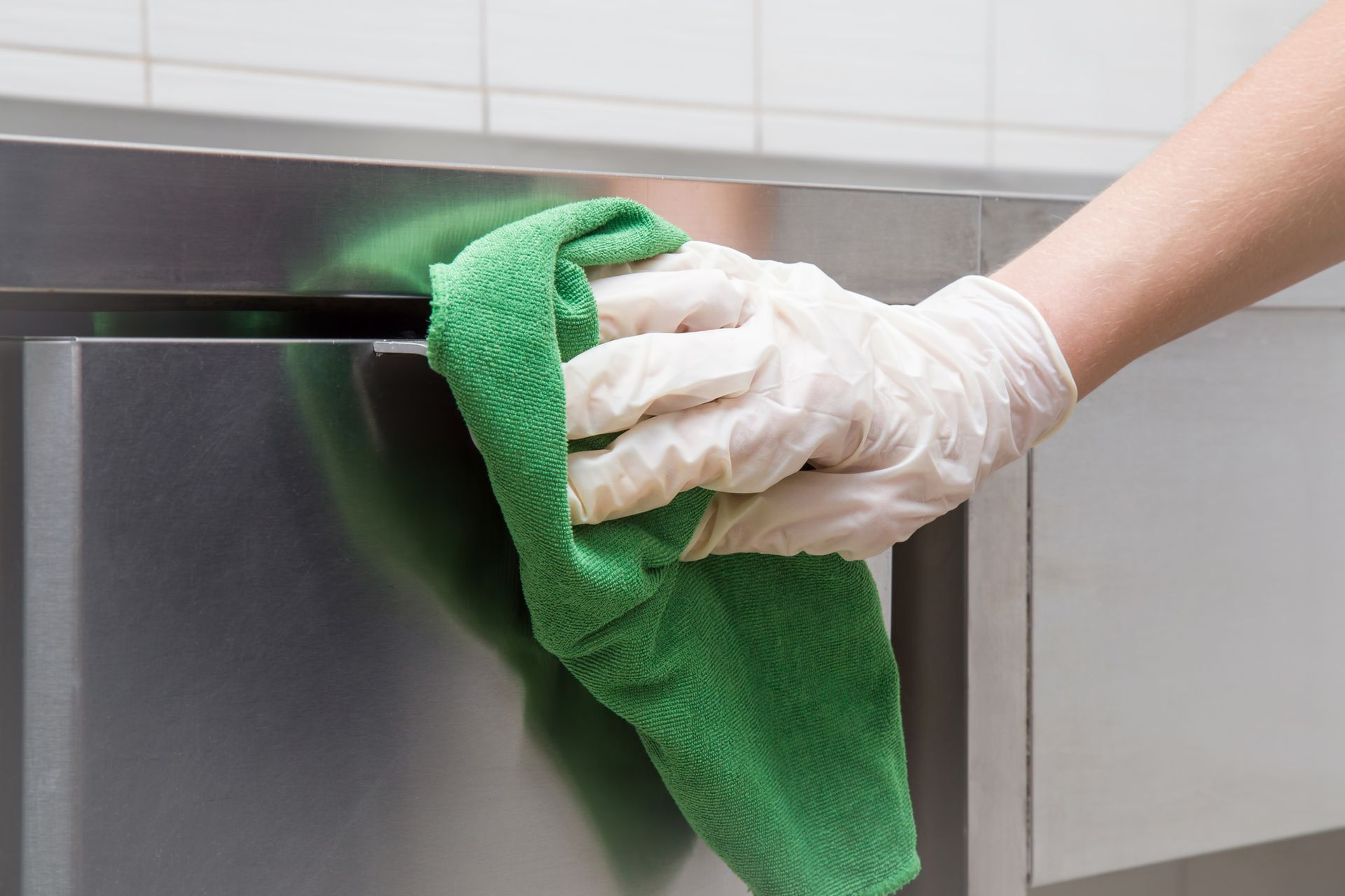 Close-up of hand in white protective gloves with green rag cleaning silver kitchen equipment.