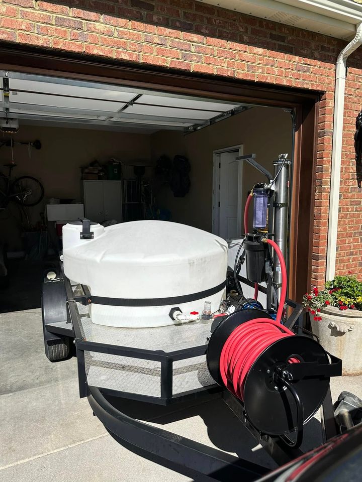 A mobile water tank trailer with a pressure washing hose reel and filtration system parked in a residential garage.