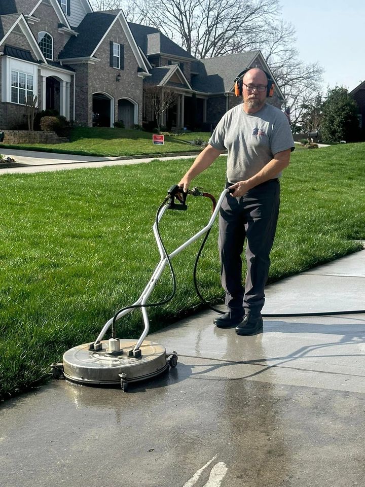 A person uses a pressure washer surface cleaner on a concrete driveway in front of a suburban house.