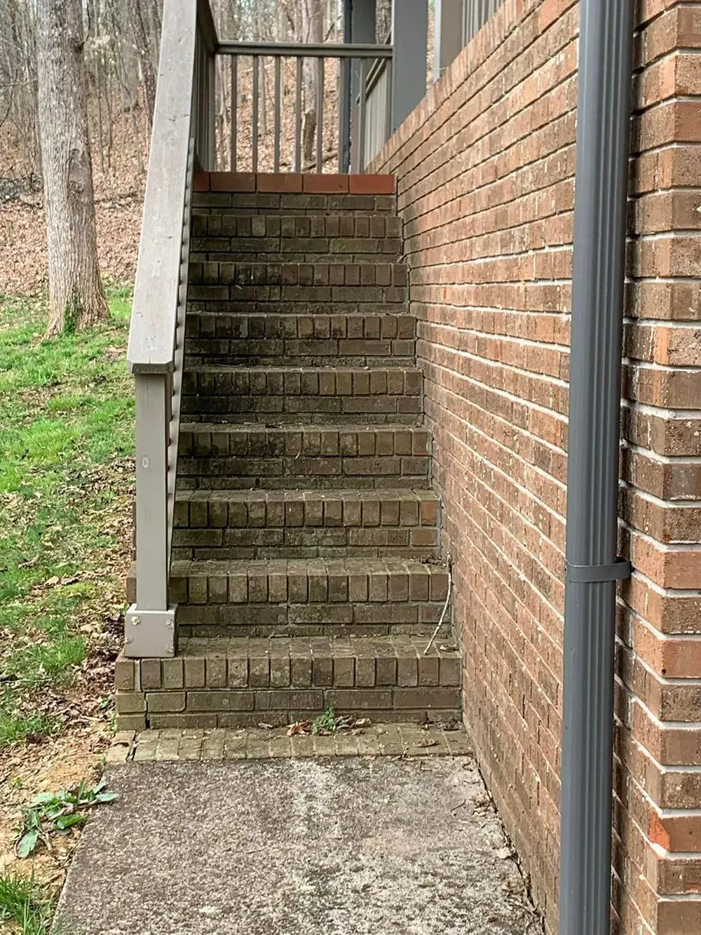 A set of outdoor brick steps with a metal railing leading up to a wooden porch beside a brick house wall.