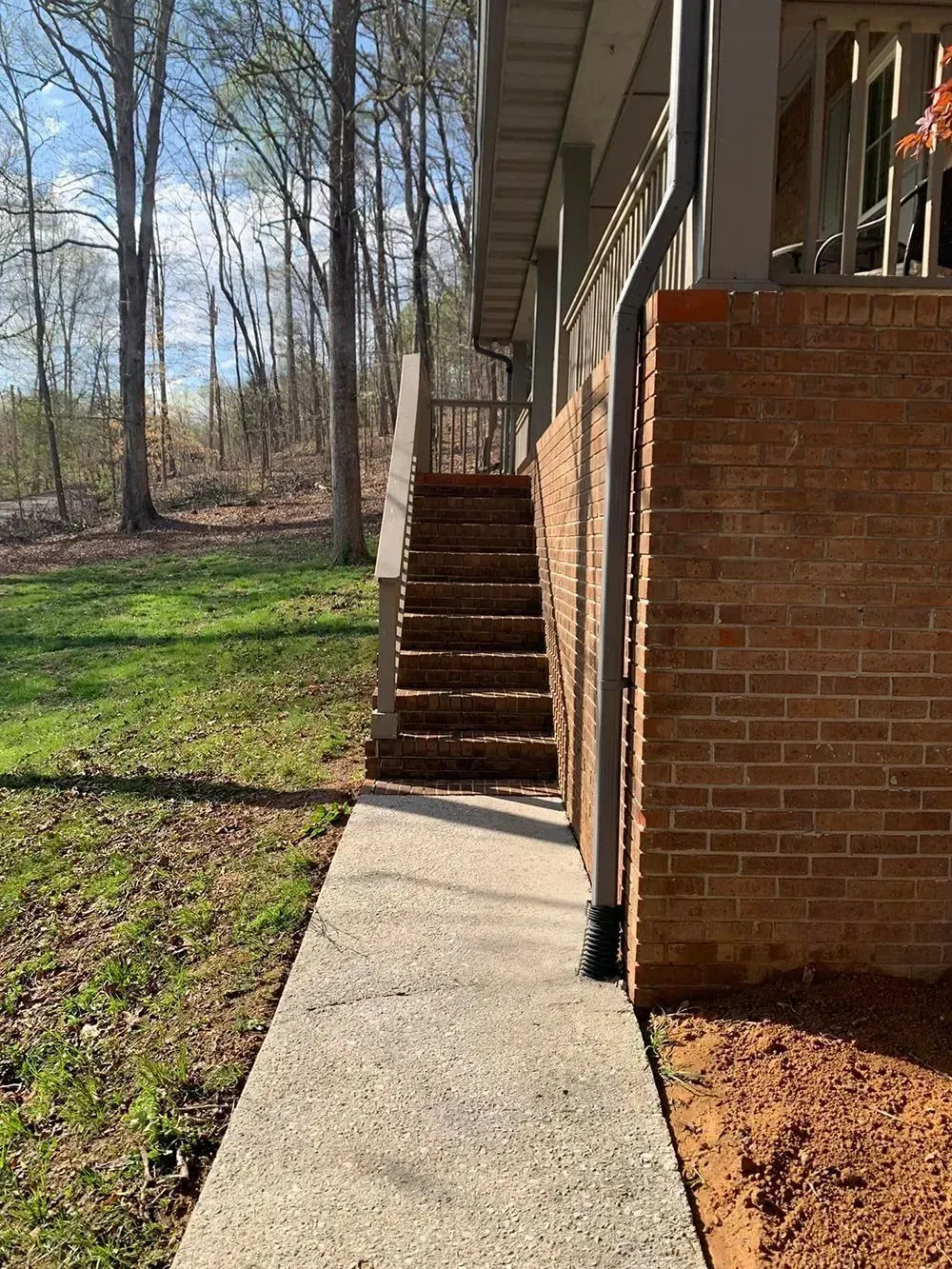 Concrete walkway leading to wooden stairs alongside a red brick wall of a house exterior near a wooded area.