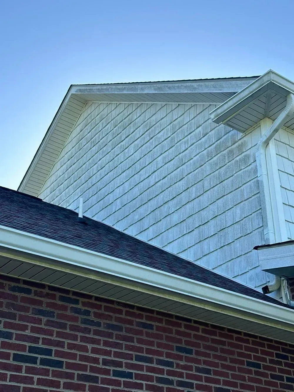 Side view of a house exterior showing light-colored vinyl siding shingles above a dark brick wall and a shingled roof.