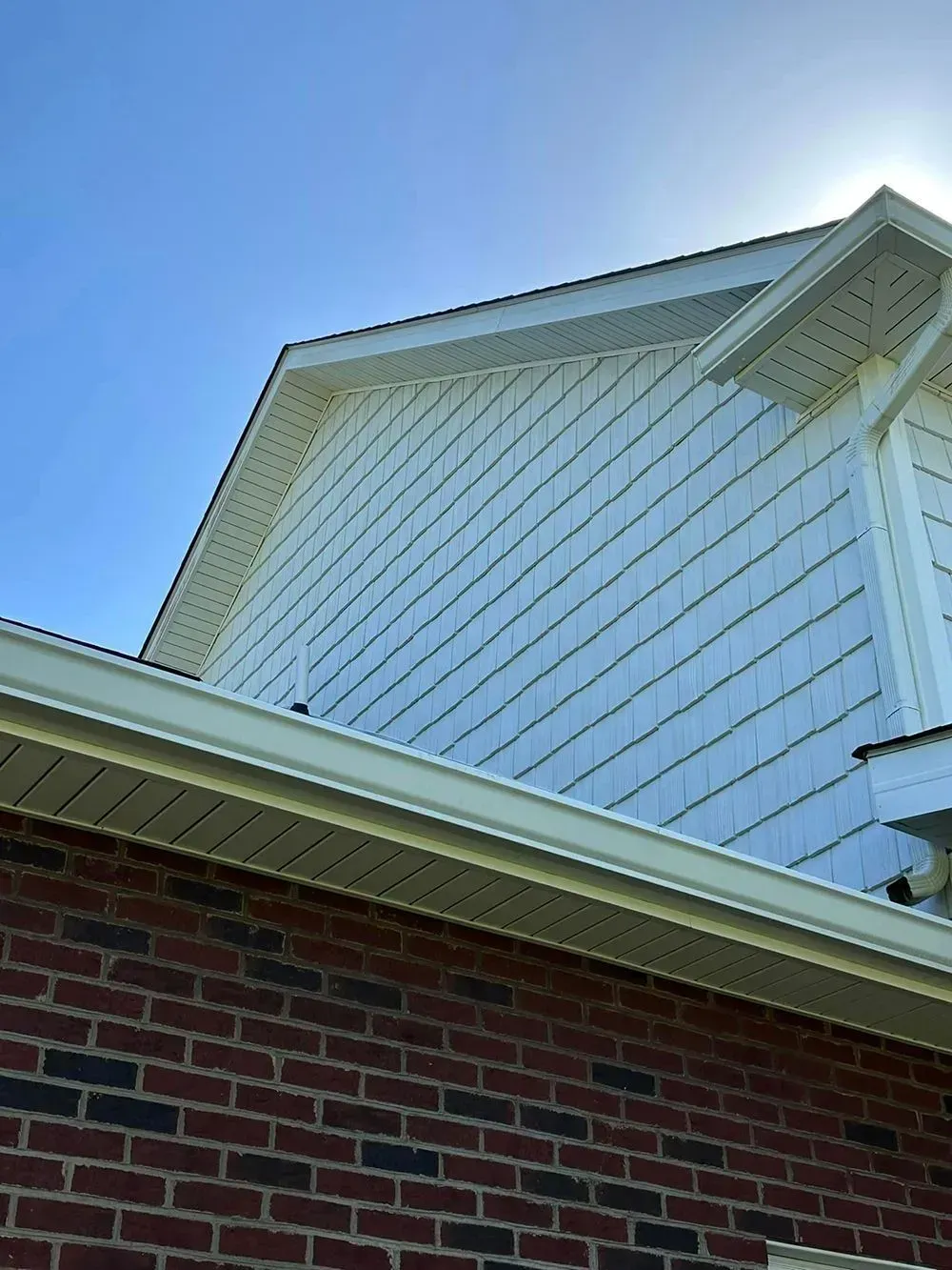 Low-angle view of a brick house wall meeting a light-colored shingle gable under a bright blue sky.