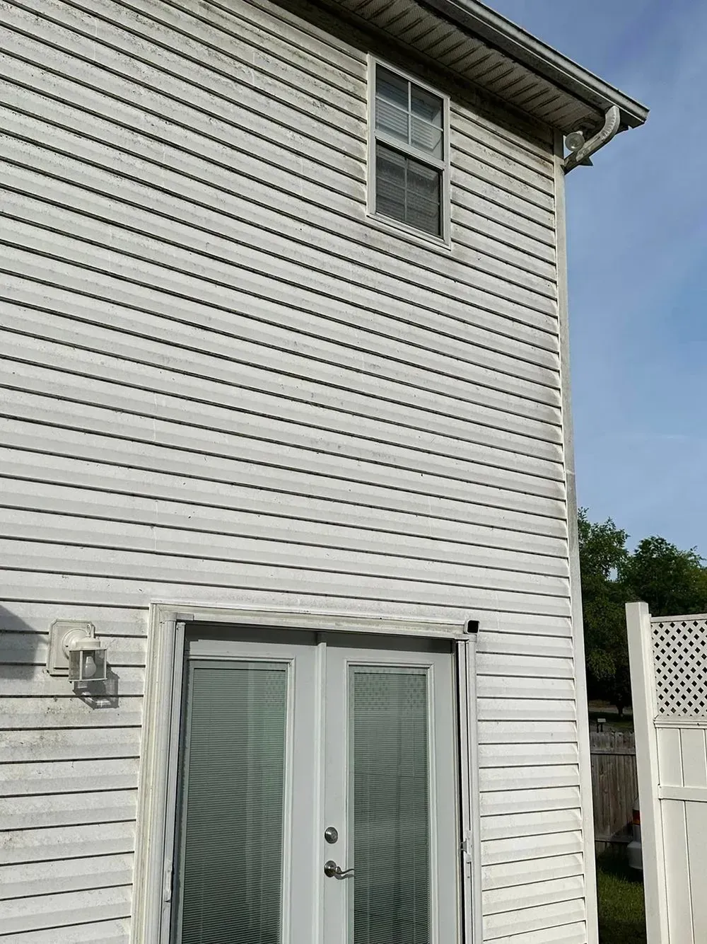 The side of a two-story house with white vinyl siding, a gray double glass door, and a single window above.